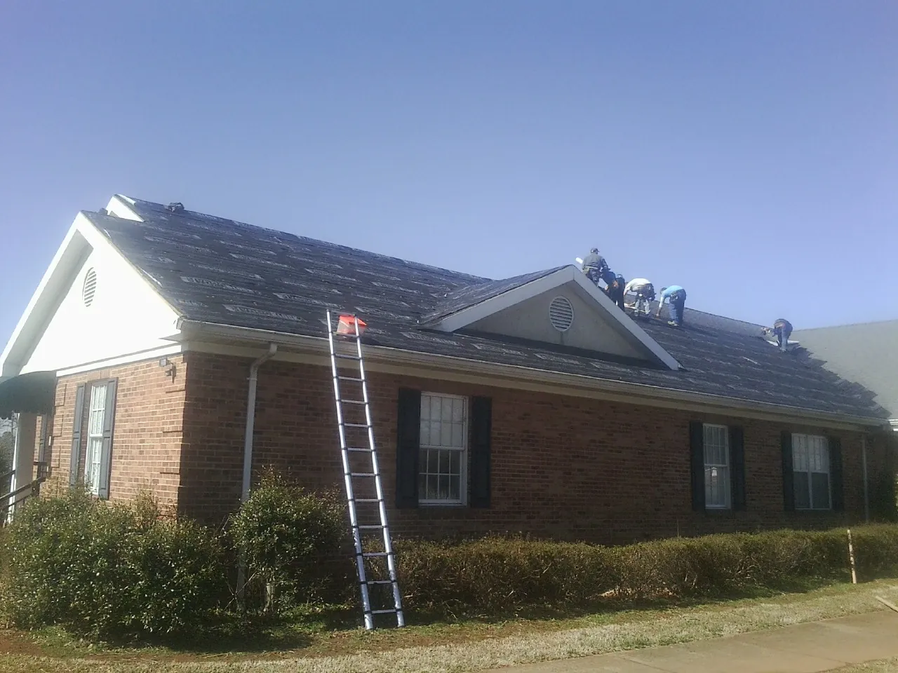 Crew tearing shingles off a brick ranch roof with a ladder set — Metal Roof Repair work by our crew in Penfield
